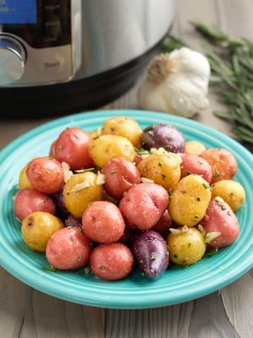 A plate of rainbow baby potatoes sprinkled with garlic and rosemary, with a sprig of rosemary, a head of garlic, and a pressure cooker in the background
