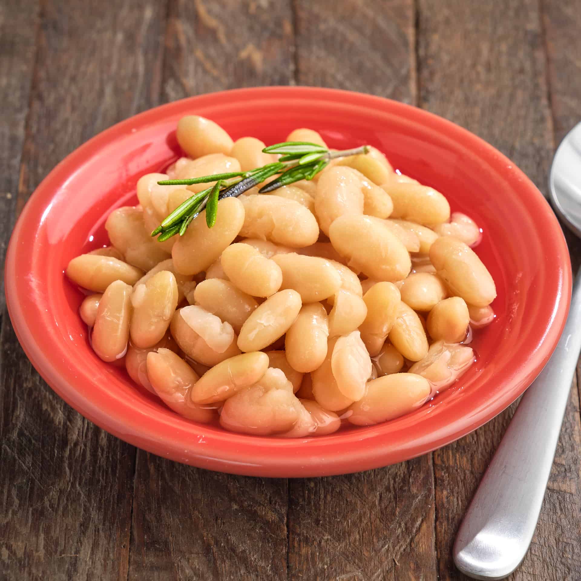 A bowl of cannellini beans with a sprig of rosemary on top and a spoon on the side