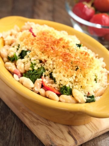 A gratin dish of beans, greens, and cherry peppers, topped with a toasted bread crumb crust, with more cherry peppers in the background