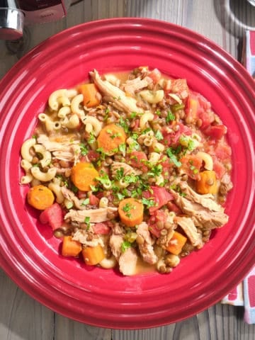 A bowl of turkey, lentil, and macaroni soup, made from the bones of a roasted turkey carcass, with a spoon, napkin, red pepper flakes, and Instant Pot in the background