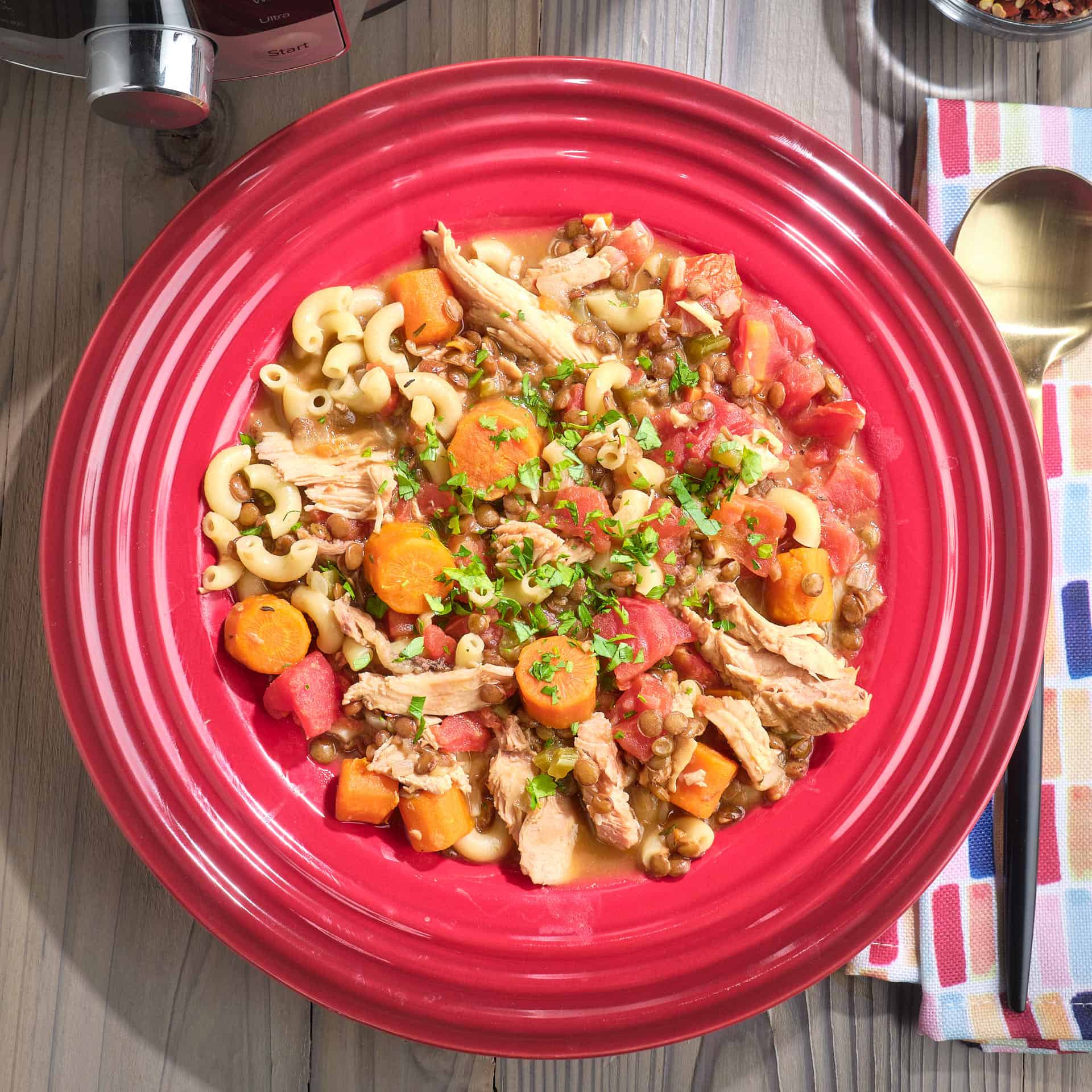 A bowl of turkey, lentil, and macaroni soup, made from the bones of a roasted turkey carcass, with a spoon, napkin, red pepper flakes, and Instant Pot in the background