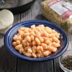 A bowl of cooked mayocoba beans, with a bag of uncooked beans, a bowl of oregano, an onion, and an Instant Pot in the background