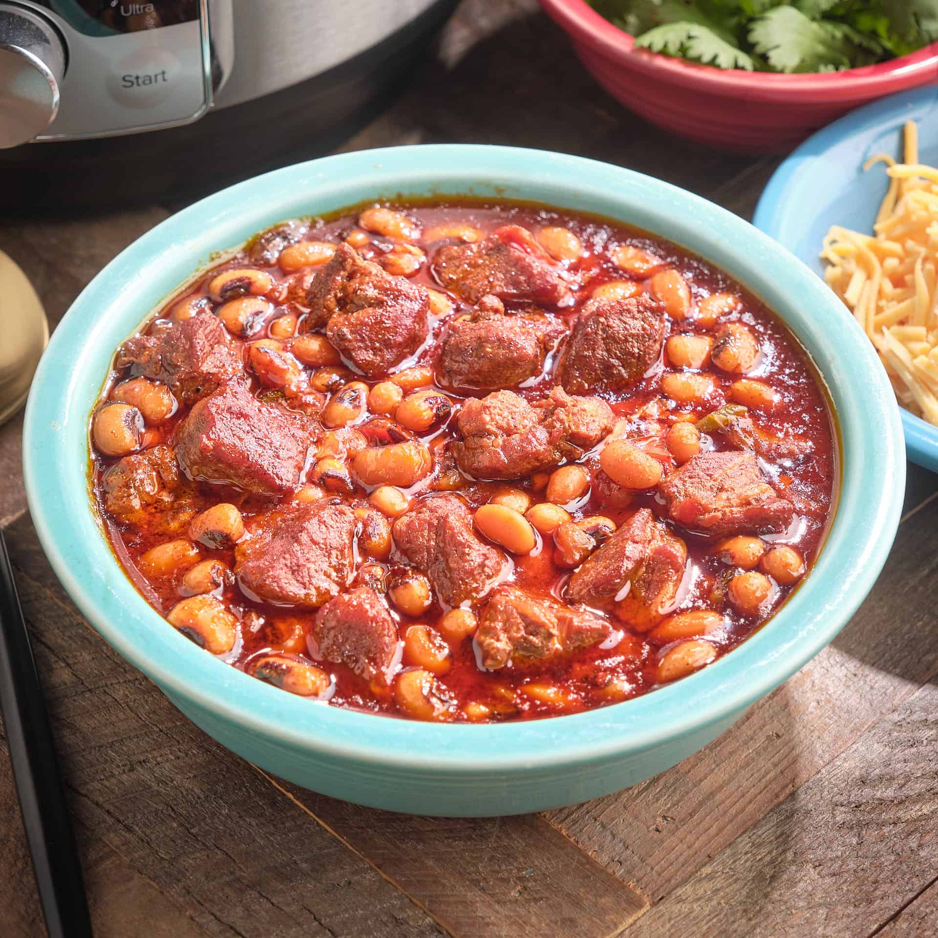 A bowl of pork and black-eyed pea chili with shredded cheese, cilantro leaves, and an Instant Pot in the background