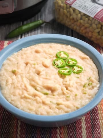 A bowl of refried mayocoba beans, with sliced serrano peppers, on a placemat next to a spoon, with uncooked beans, a serrano pepper, and an Instant Pot in the background