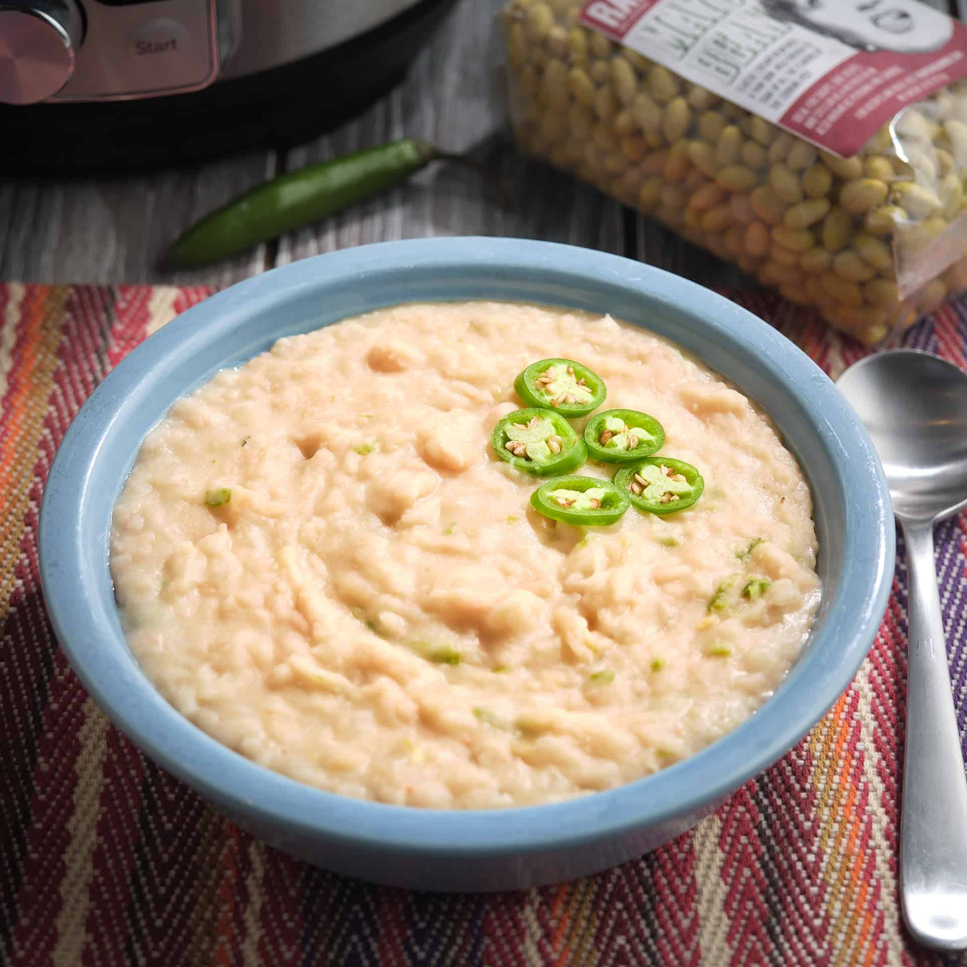 A bowl of refried mayocoba beans, with sliced serrano peppers, on a placemat next to a spoon, with uncooked beans, a serrano pepper, and an Instant Pot in the background