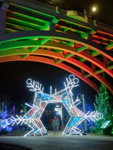 Lit up star with a woman standing inside, with the Fulton Road Bridge in the background, lit up with holiday colors