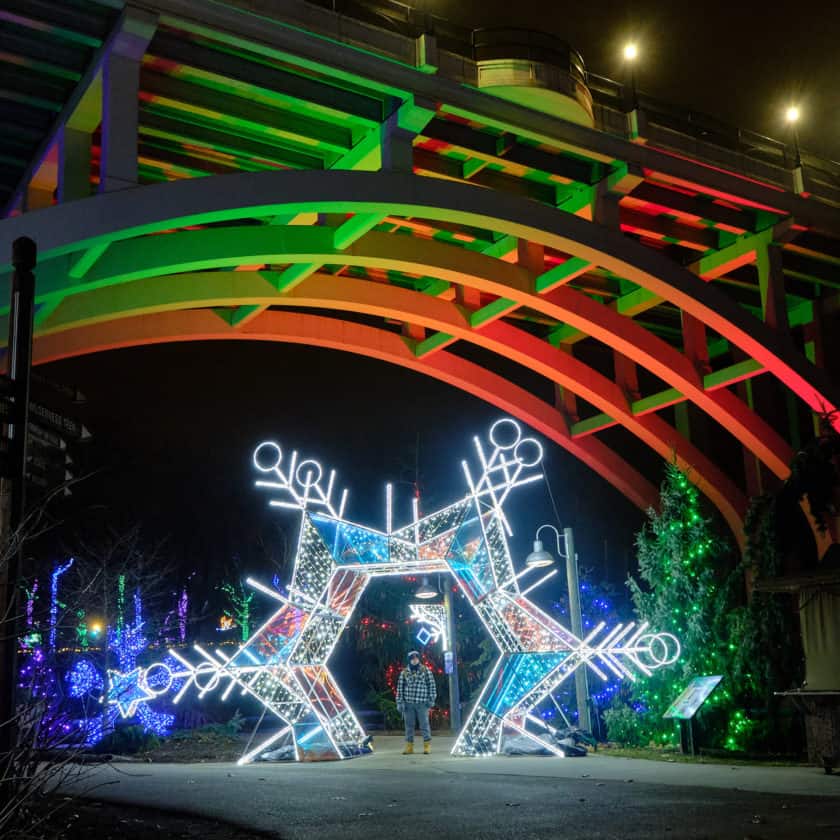 Lit up star with a woman standing inside, with the Fulton Road Bridge in the background, lit up with holiday colors