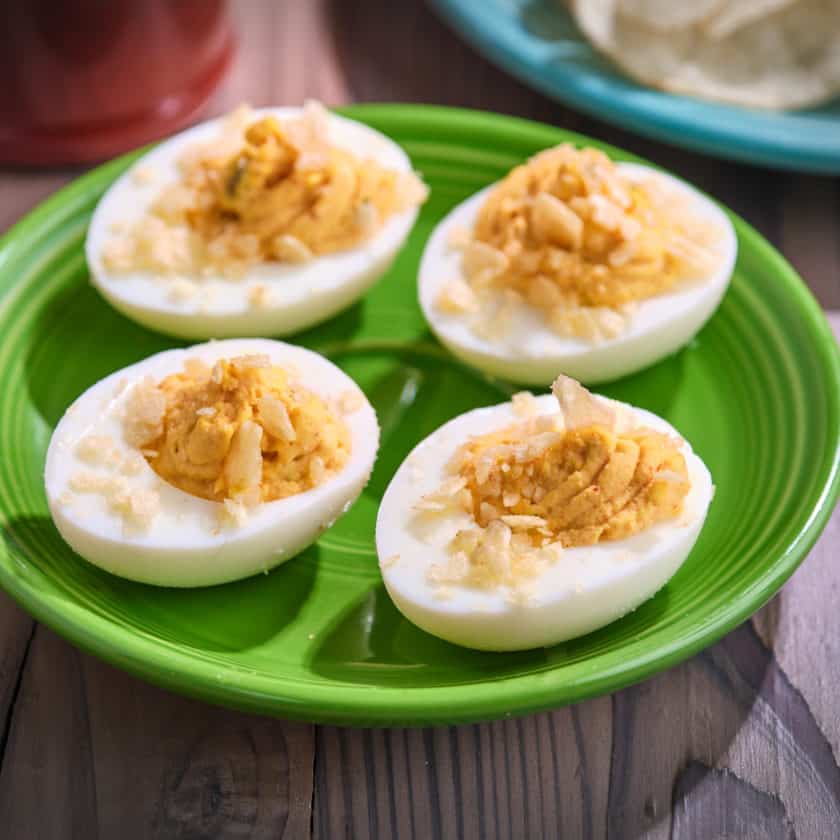 A plate of deviled eggs topped with crushed potato chips, with more potato chips and some paprika in the background