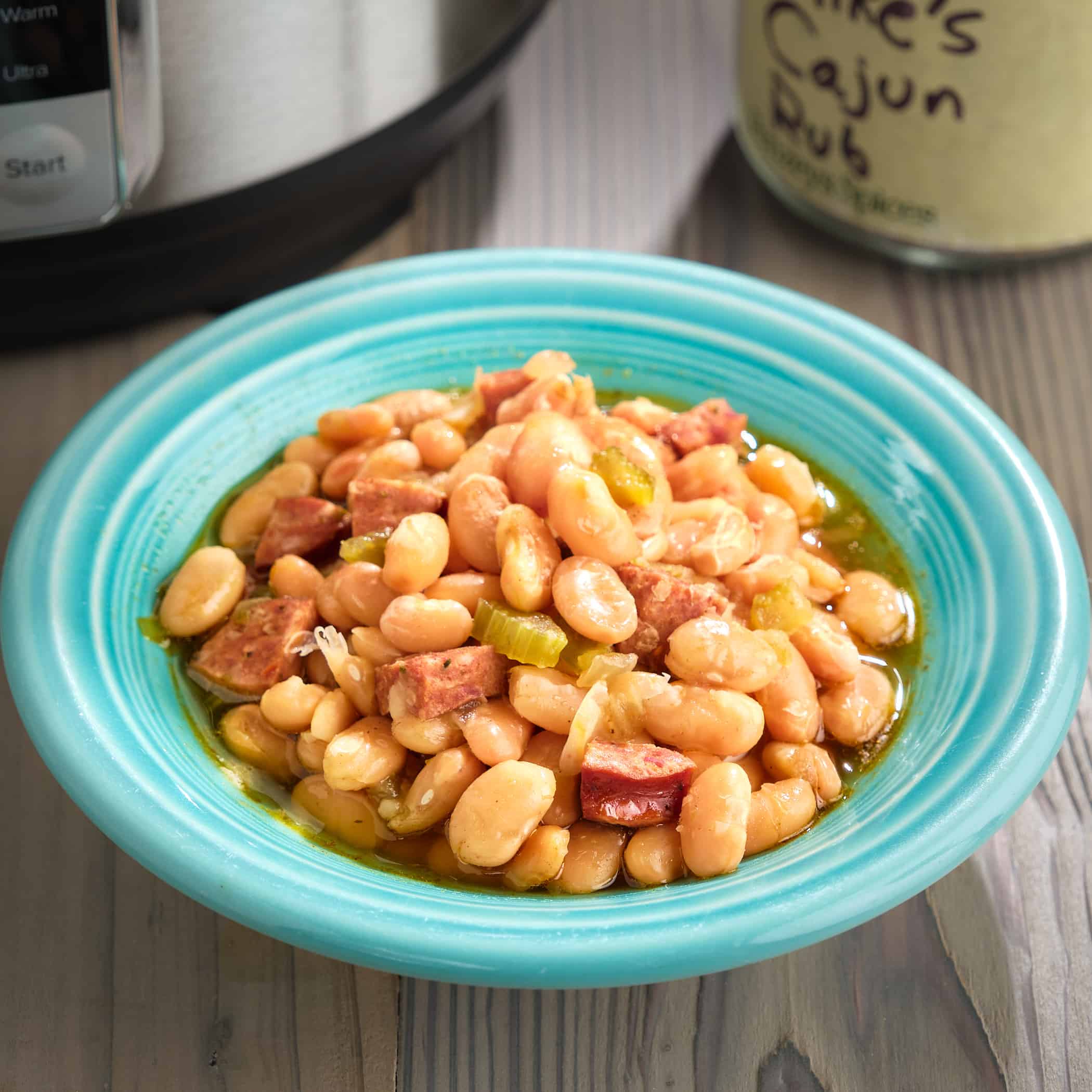 A bowl of cajun white beans, with sausage and green peppers, in front of an Instant Pot and a jar of Cajun seasoning