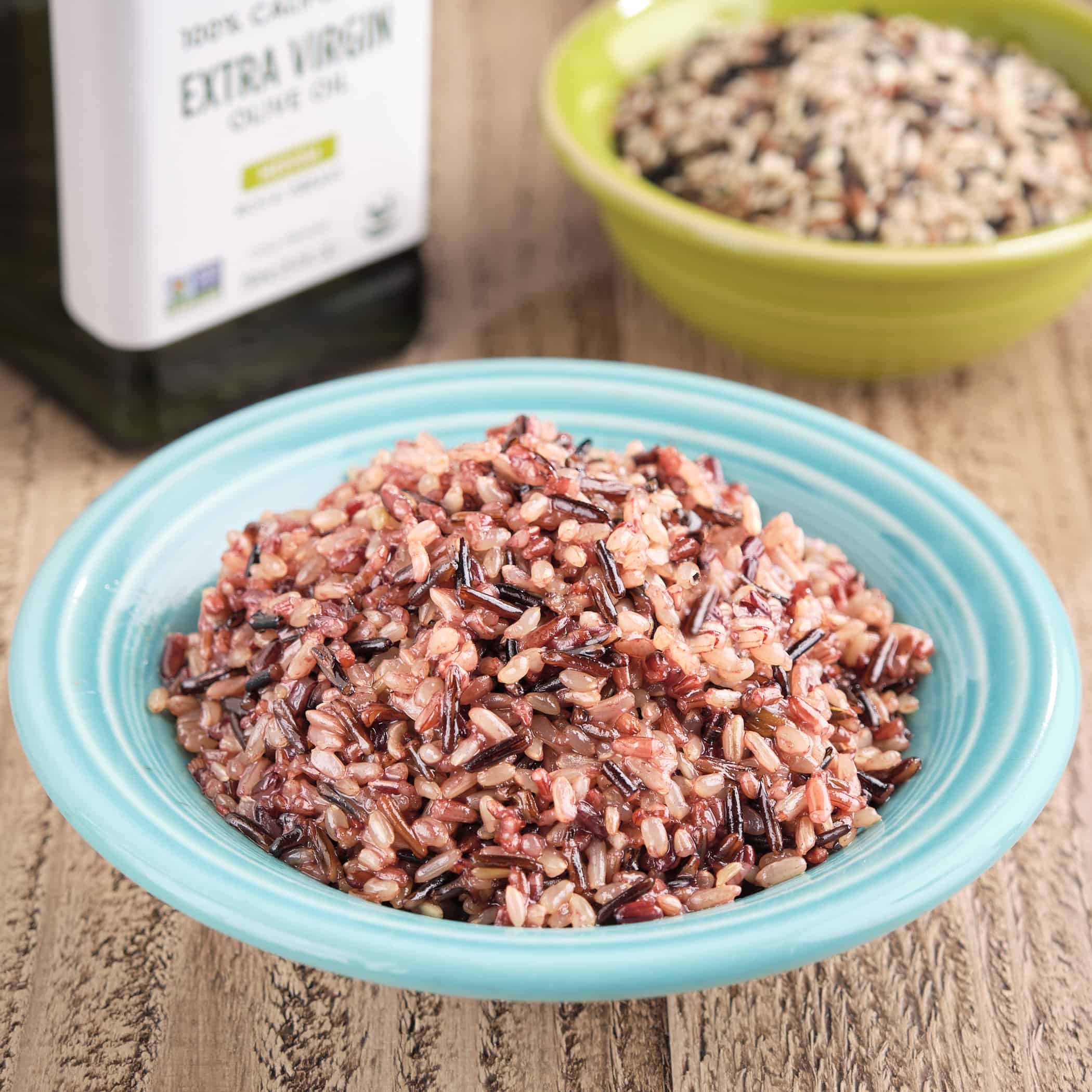 A bowl of wild rice blend, with an uncooked bowl of rice and a bottle of olive oil in the background