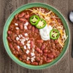 A bowl of ground beef and bean chili, topped with diced onion, sour cream, jalapenos, and shredded cheese
