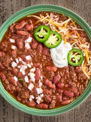 A bowl of ground beef and bean chili, topped with diced onion, sour cream, jalapenos, and shredded cheese
