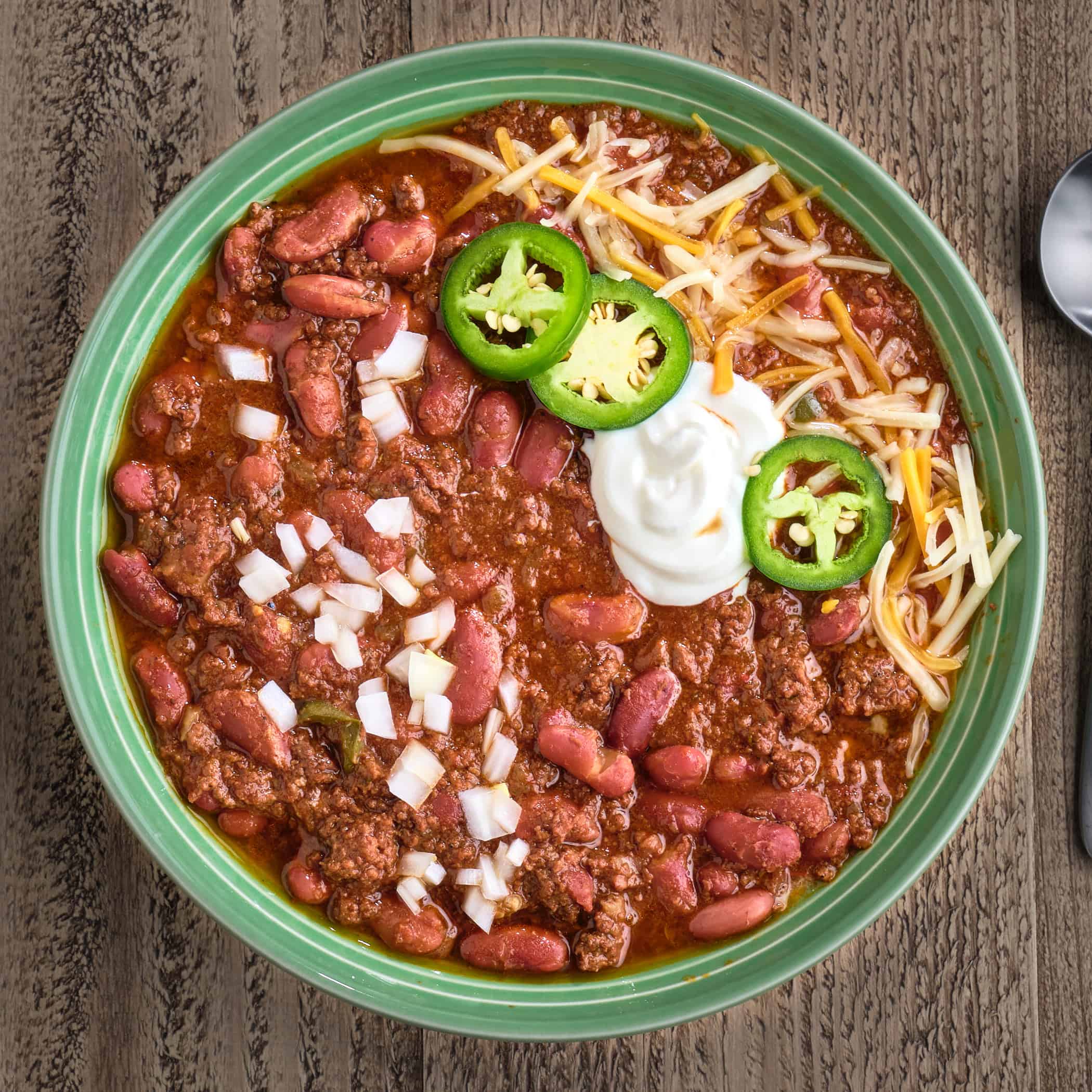 A bowl of ground beef and bean chili, topped with diced onion, sour cream, jalapenos, and shredded cheese