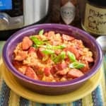 A bowl of brown rice jambalaya with sausage, chicken, and green onions, and tabasco sauce and cajun rub in the background.