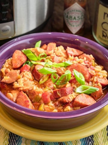 A bowl of brown rice jambalaya with sausage, chicken, and green onions, and tabasco sauce and cajun rub in the background.