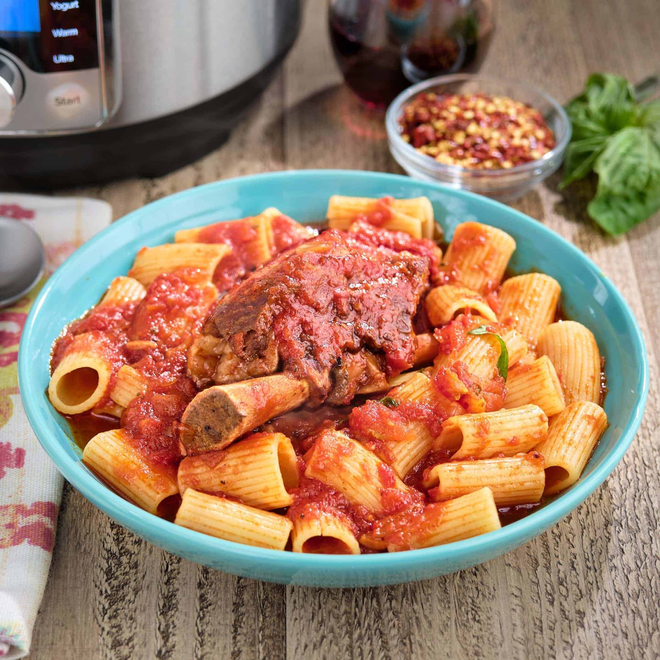 A bowl of pasta, sauced with ragu Tucci, topped with a big beef short rib, with an Instant Pot, a glass of wine, and a bowl of red pepper flakes in the background.