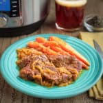 A plate of chuck steaks with beer and onions, carrots, and a glass of beer in the background.
