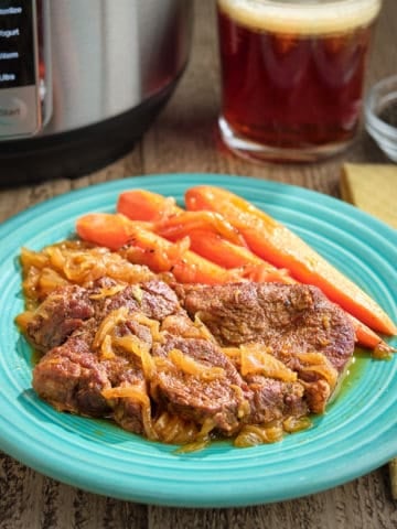 A plate of chuck steaks with beer and onions, carrots, and a glass of beer in the background.