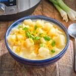 A bow of potato soup topped with shredded cheddar and sliced green onion, on a wooden table with an Instant Pot in the background.