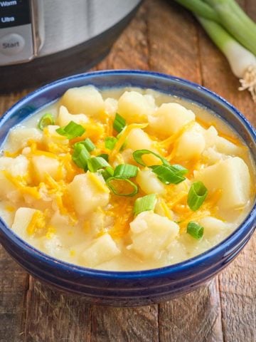 A bow of potato soup topped with shredded cheddar and sliced green onion, on a wooden table with an Instant Pot in the background.