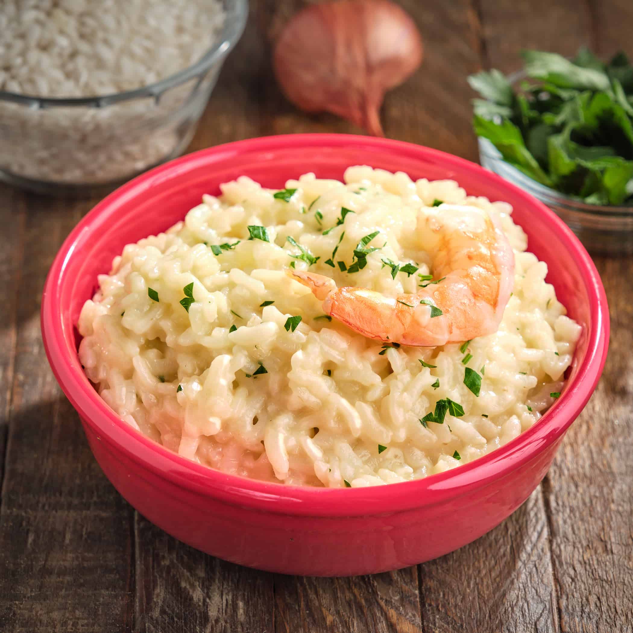 A bowl of shrimp risotto, with parsley, shallot, and uncooked rice in the background.