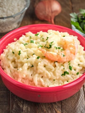 A bowl of shrimp risotto, with parsley, shallot, and uncooked rice in the background.