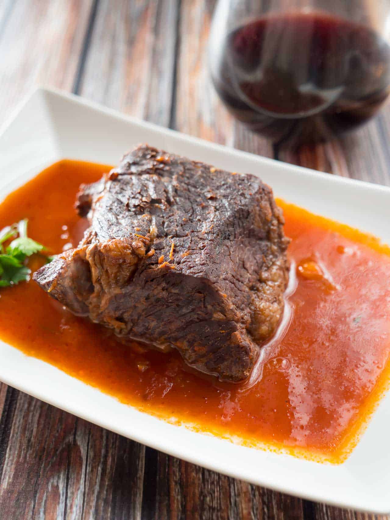 A piece of pot roast surrounded by tomato sauce on a white plate with a glass of wine in the background