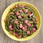 A yellow bowl of cooked turnip greens and ham on a wood table
