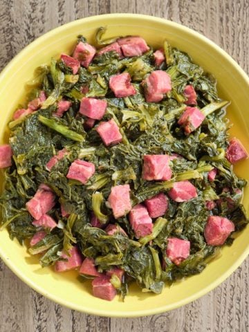 A yellow bowl of cooked turnip greens and ham on a wood table