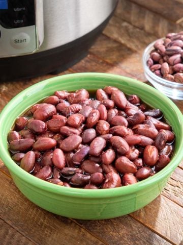 A bowl of cooked Rio Zape beans on a wood table, with a dish of uncooked beans and an Instant Pot in the background