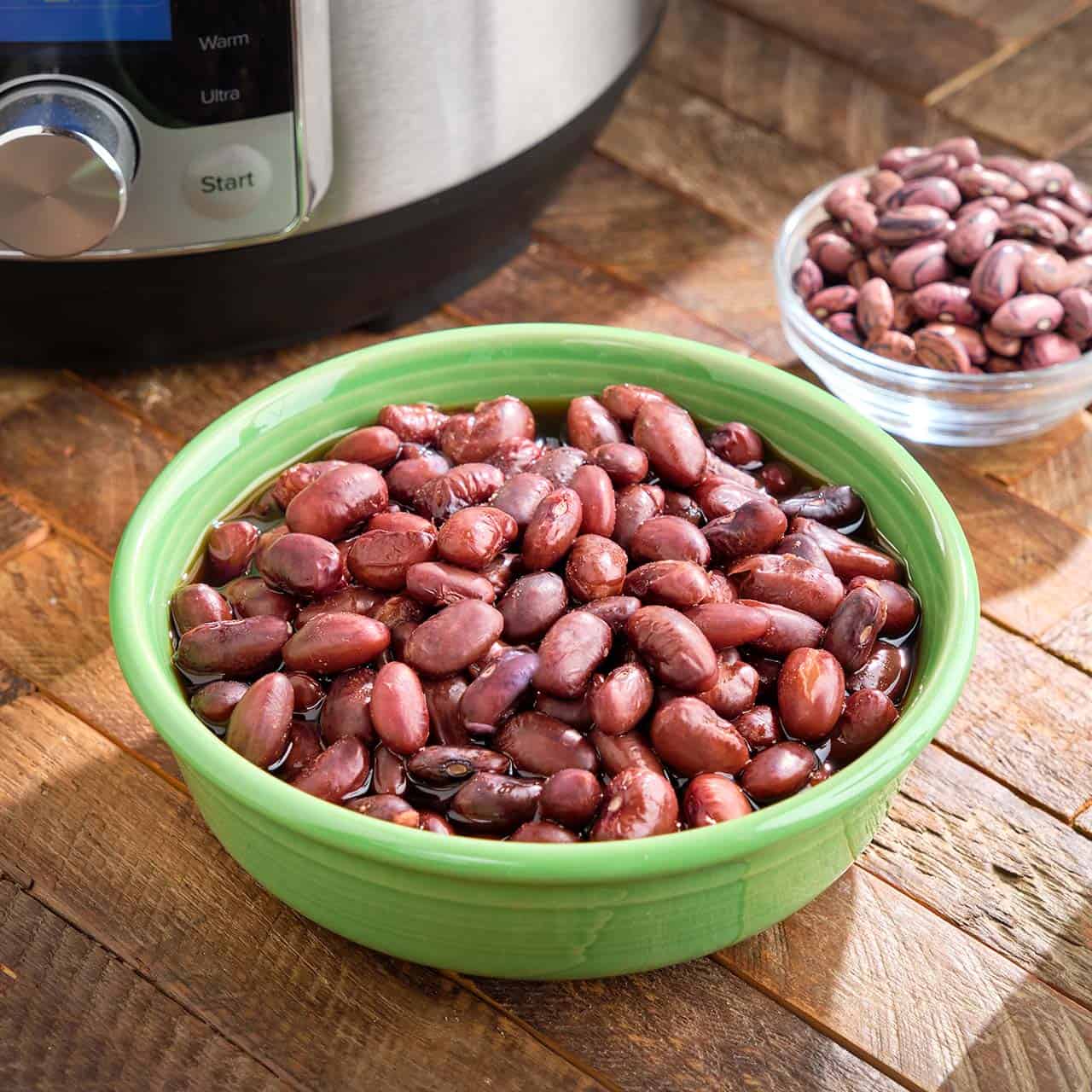 A bowl of cooked Rio Zape beans on a wood table, with a dish of uncooked beans and an Instant Pot in the background