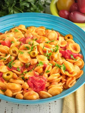 A bowl of chickpea puttanesca, with napkins, a spoon, cherry tomatoes, and parsley in the background.