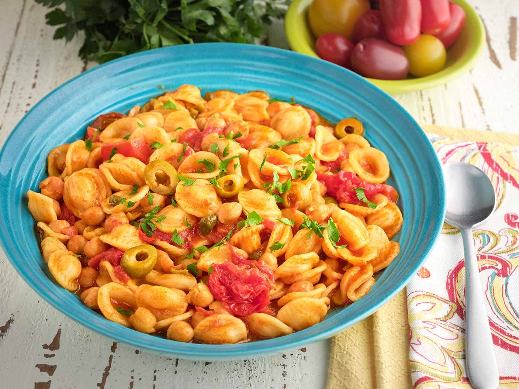 A bowl of chickpea puttanesca, with napkins, a spoon, cherry tomatoes, and parsley in the background.