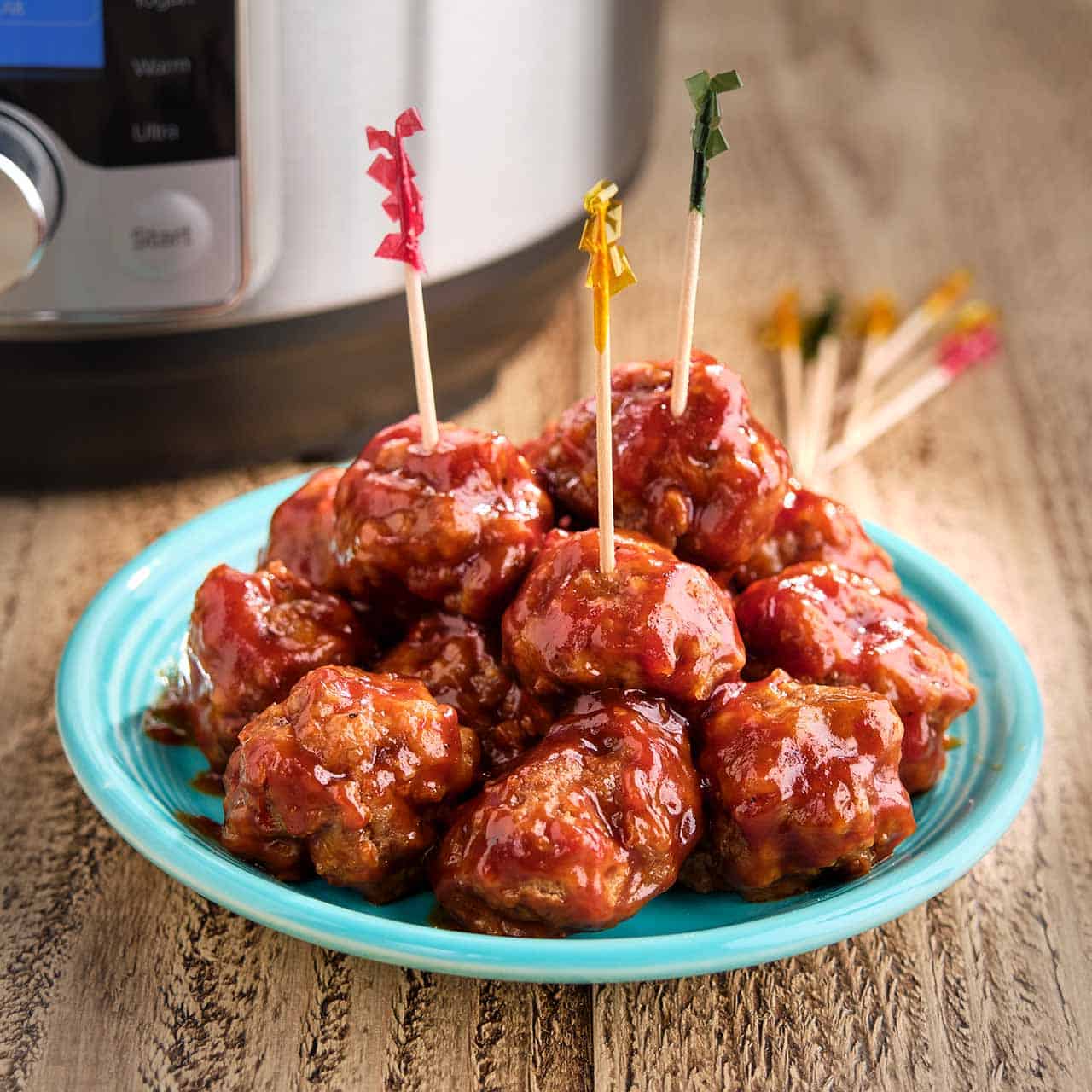 A plate of BBQ meatballs with frilly toothpicks, and an Instant Pot and more toothpicks in the background