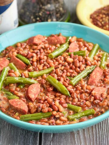 A bowl of field peas and snapped green beans, with red pepper flakes, black pepper, and hot sauce in the background