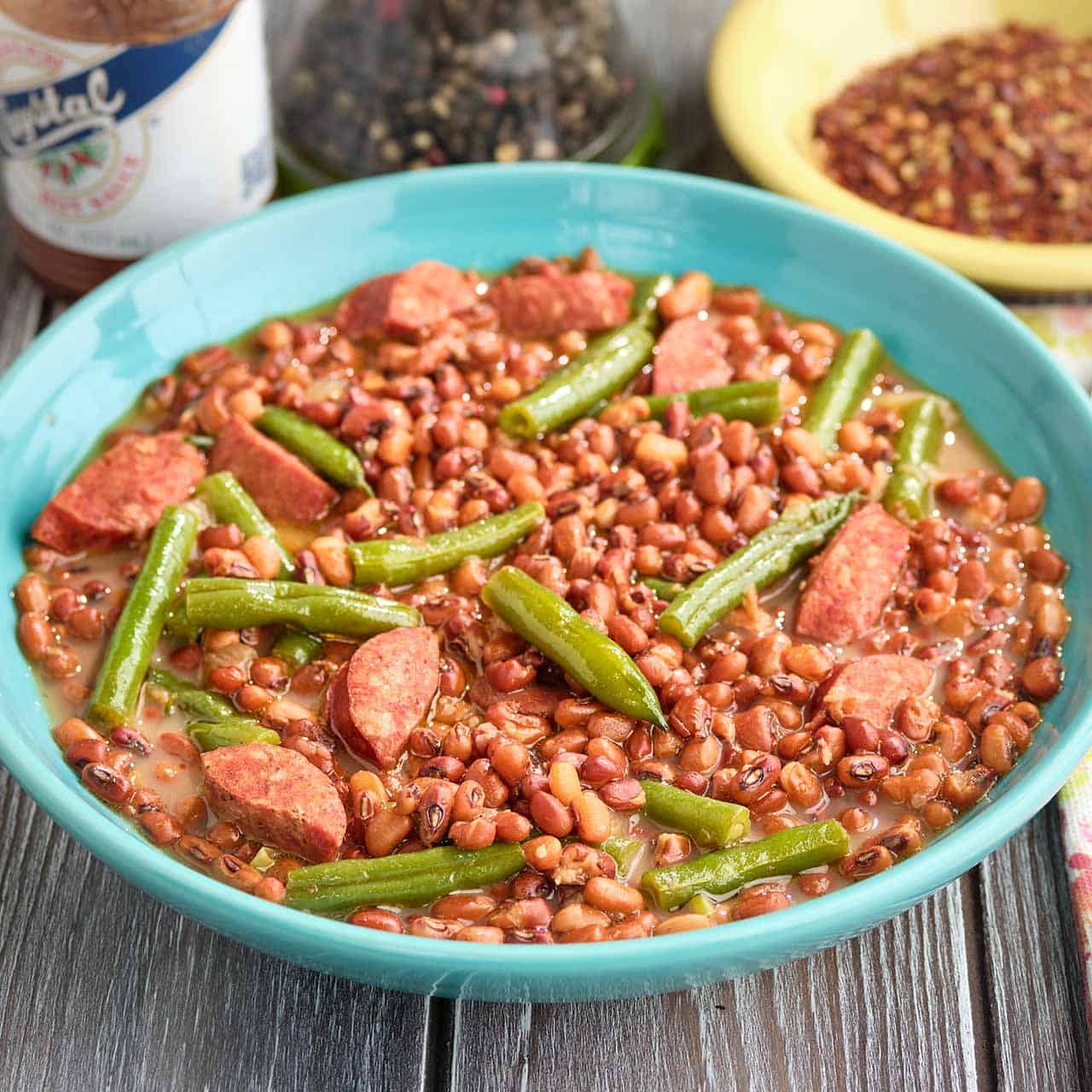 A bowl of field peas and snapped green beans, with red pepper flakes, black pepper, and hot sauce in the background