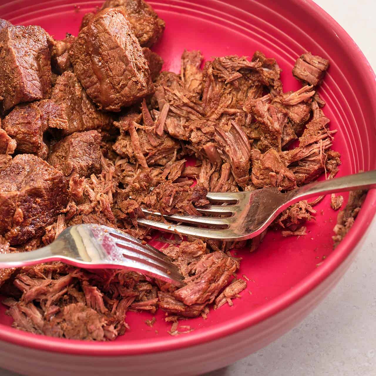 A bowl with cubes of beef, a pair of forks, and some shredded beef