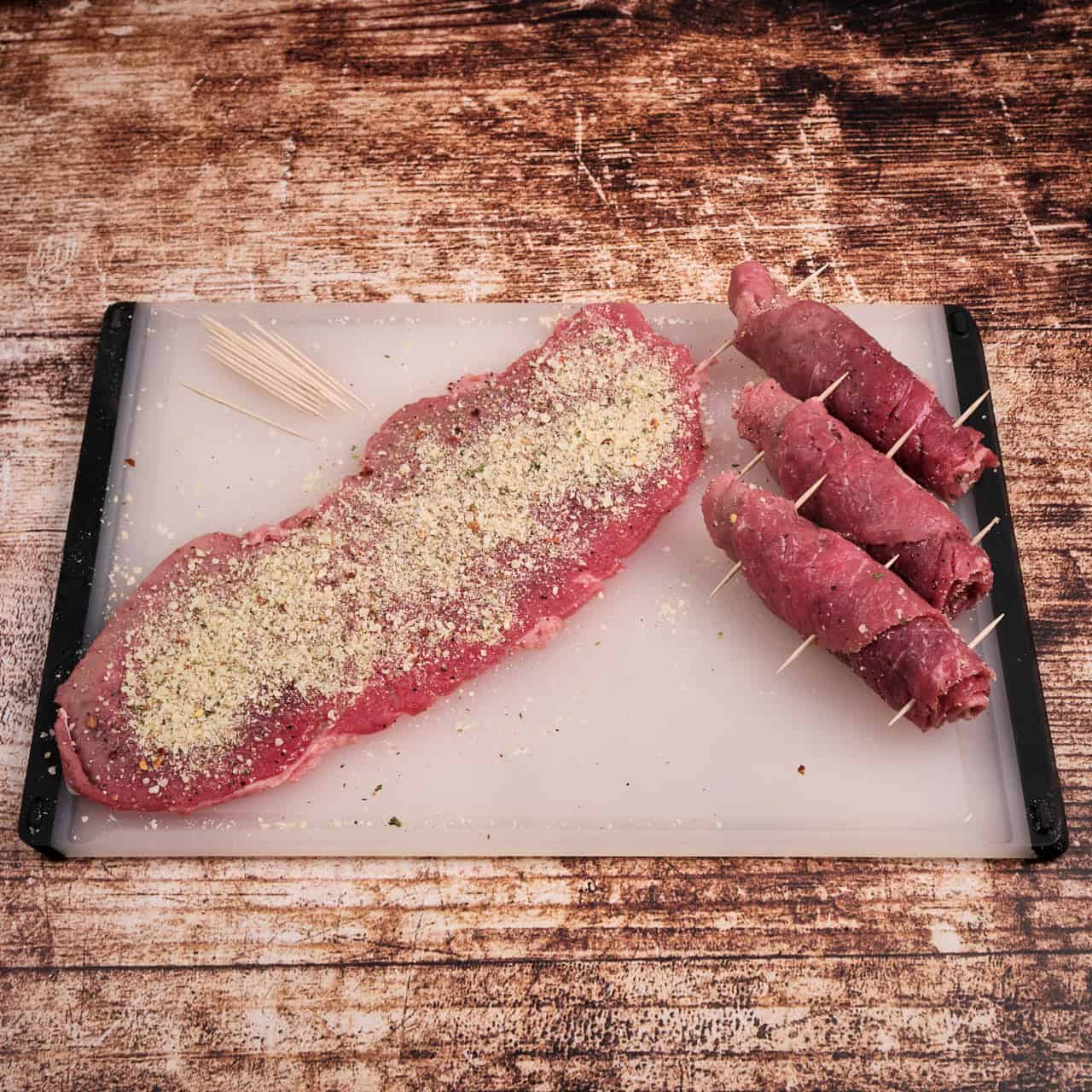 A slice of beef sprinkled with the filling, with toothpicks and a few beef rolls next to it