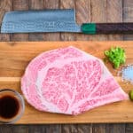 A Wagyu ribeye steak on a cutting board with wasabi, ponzu sauce, salt, green onions, and a knife.