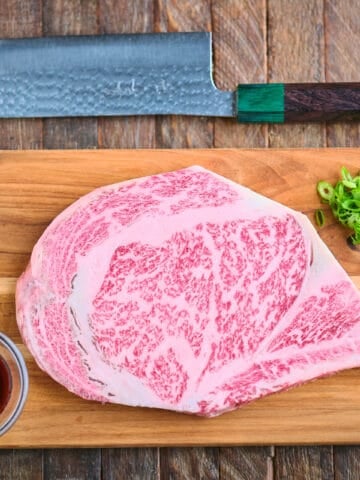 A Wagyu ribeye steak on a cutting board with wasabi, ponzu sauce, salt, green onions, and a knife.
