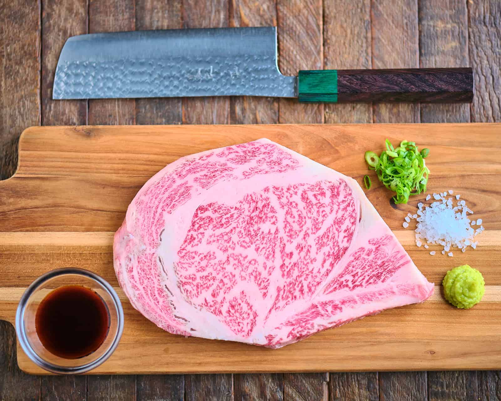 A Wagyu ribeye steak on a cutting board with wasabi, ponzu sauce, salt, green onions, and a knife.