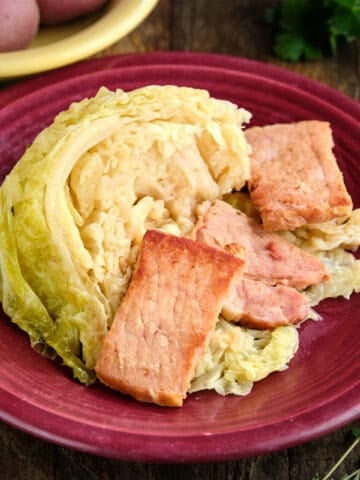 A plate of instant pot cabbage with bacon and butter, with herbs and potatoes in the background