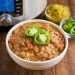 A bowl of Chili Verde with ground pork, and an Instant Pot, sliced Jalapenos, and diced chiles in the background