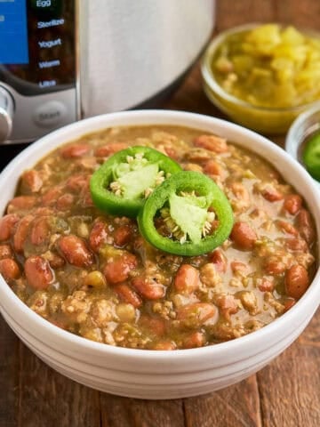 A bowl of Chili Verde with ground pork, and an Instant Pot, sliced Jalapenos, and diced chiles in the background