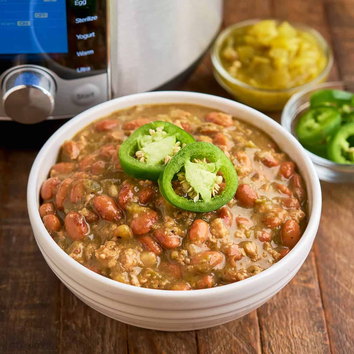 A bowl of Chili Verde with ground pork, and an Instant Pot, sliced Jalapenos, and diced chiles in the background