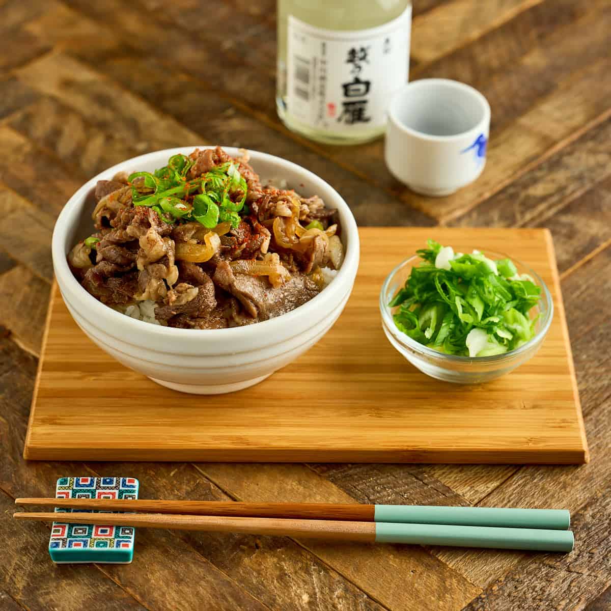 A bowl of gyudon with extra sliced green onions, chopsticks, and sake